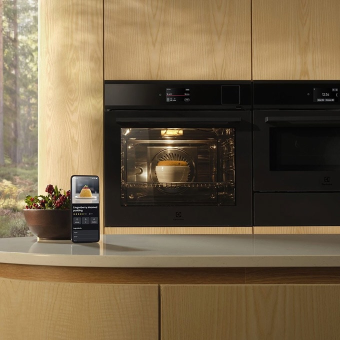 Två svarta Electrolux-ugnar inbyggda i ljust trä, där en smartphone visar AI TasteAssist-appen