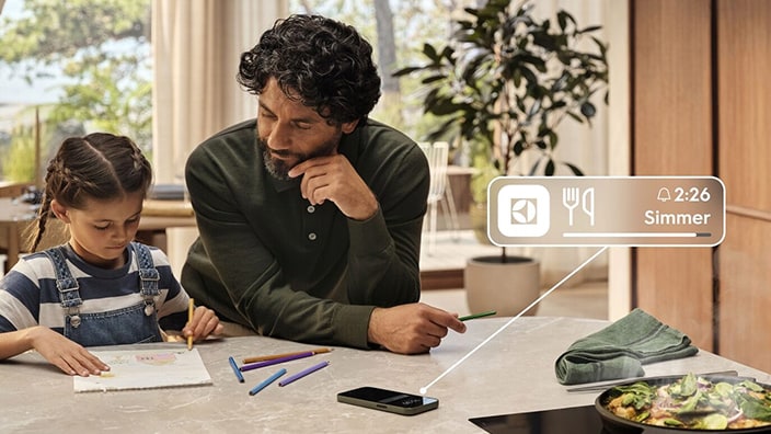 En pappa och dotter ritar vid köksbordet medan en smartphone visar en notis från Electrolux-appen