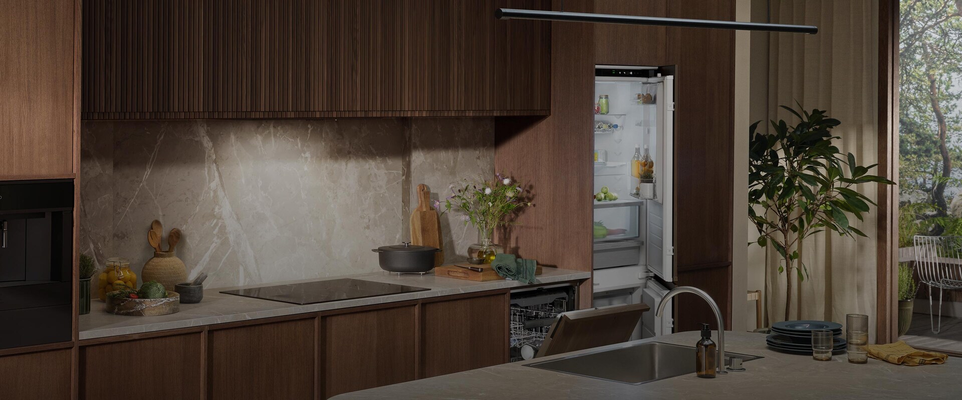 Integrerad kyl-frys och diskmaskin i ett stilrent kök med mörka träpaneler och bänkskiva i marmor.