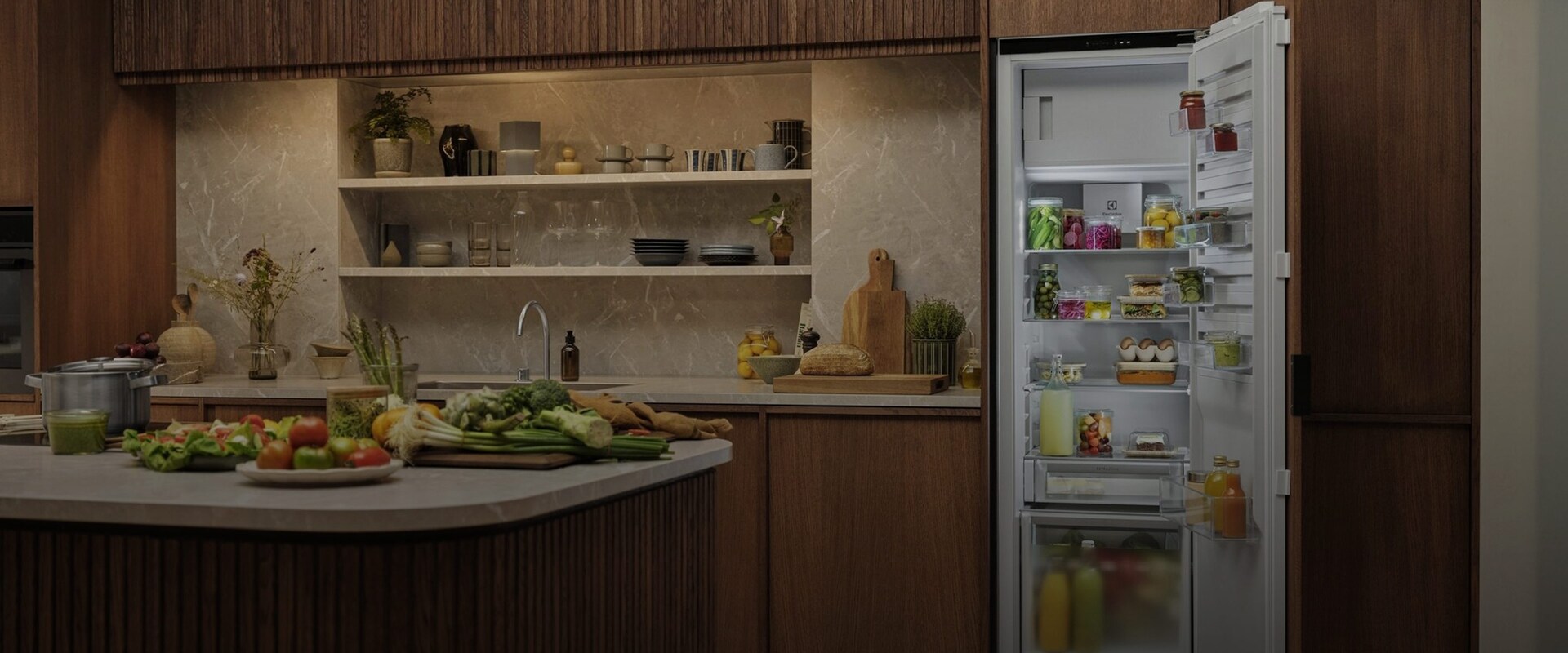 En överblick av ett öppet integrerat Electrolux kylskåp i ett modernt kök, fyllt med färska råvaror, flaskor och organiserade matlådor.